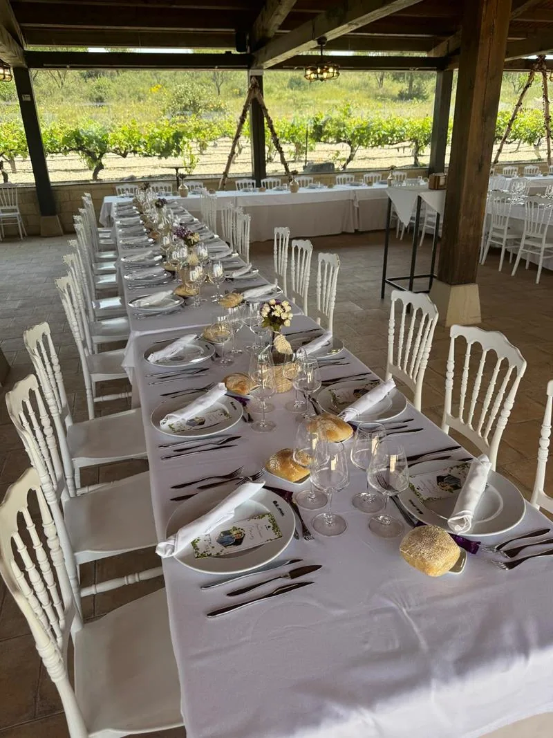 Mesa larga vestida con sillas blancas y cristalería, junto al viñedo